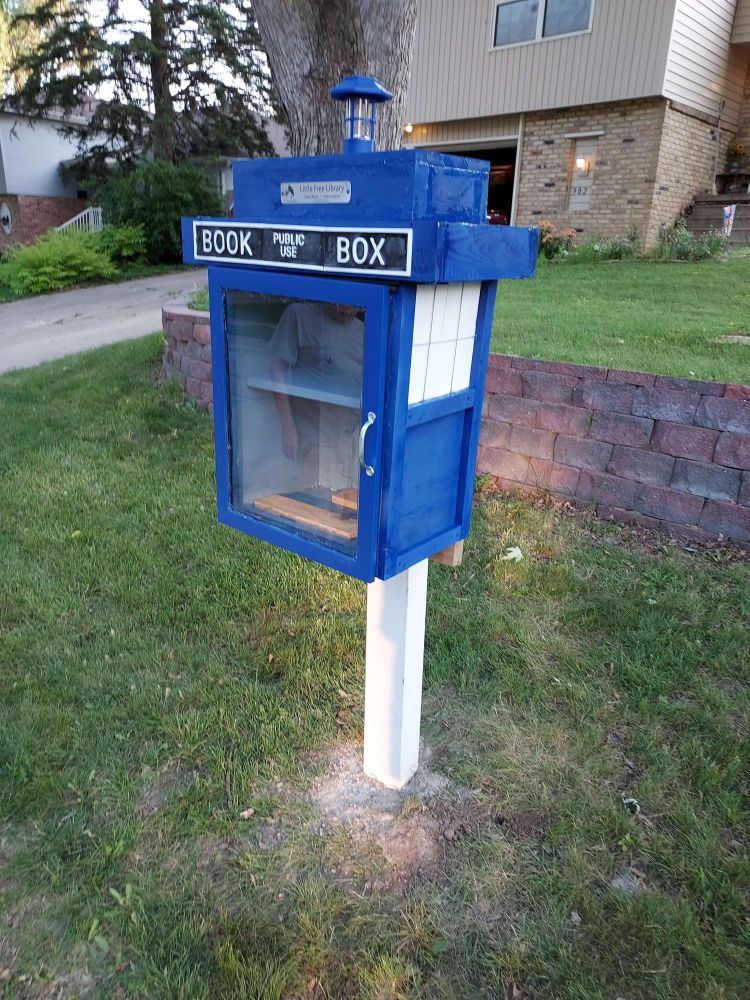 The little free library TARDIS installed! The blue box is hung up on a white post in front of a stone wall.