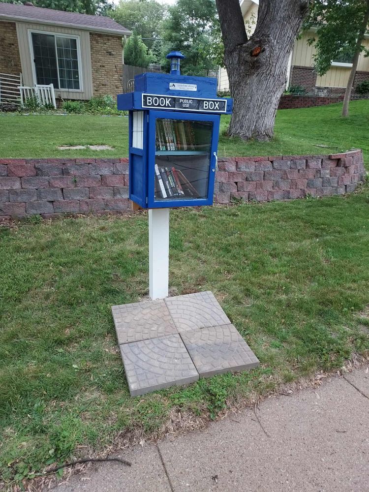 a blue Little Free Library designed to look like a TARDIS from Doctor Who, mounted on a pole. It's over a group of paving stones and in front of a landscaping wall.