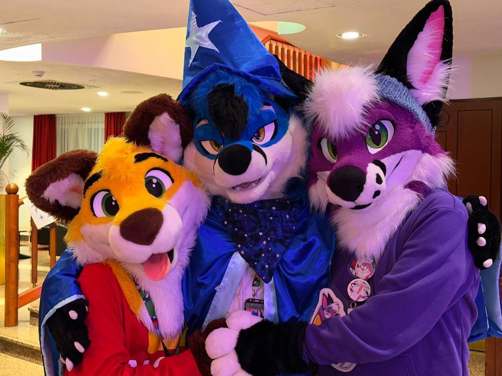Nico, Blujay and Fizz (yellow, blue and purple foxes) posing at Awoostria, hugging each other. Blujay is wearing a blue wizard hat and garb. Nico and Fizz are wearing red and purple sweaters respectively. Photo by @coorandilla.bsky.social