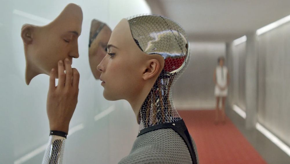 Still from EX MACHINA. A female-presenting android contemplates the realistic face-mask of a different android, in a brightly lit hallway with red carpeting. Another android figure stands in the background. 