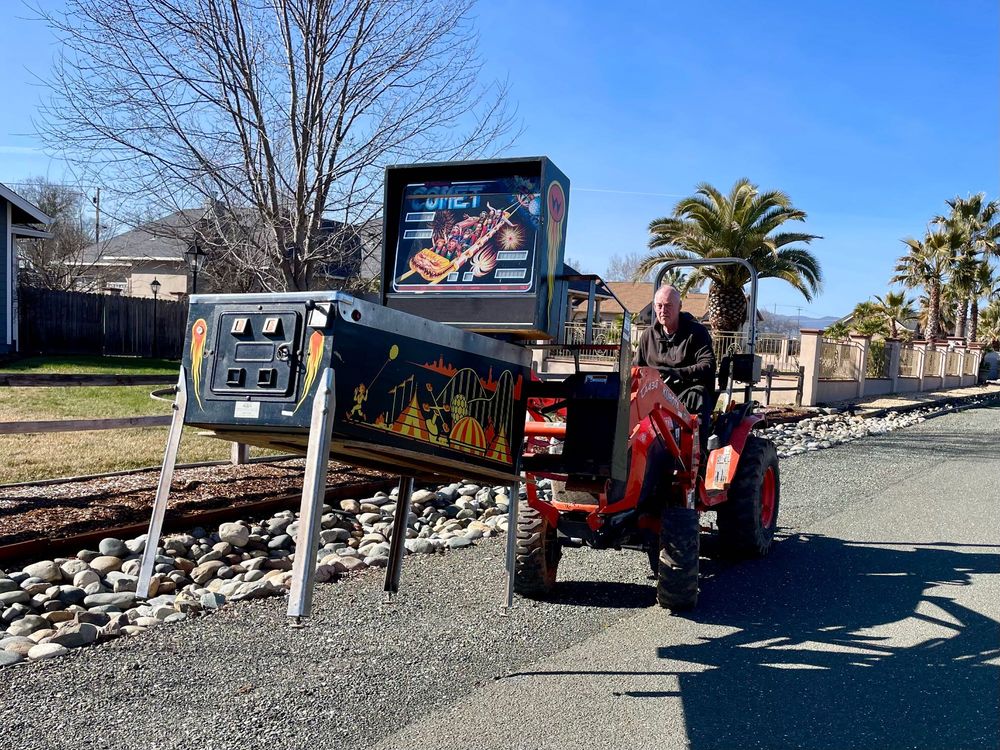 Pinball machine being lifted by a tractor with forks on the front. The backbox has not been folded down.

This is not how you should transport a pinball machine. This is my father-in-law doing things his way because that's how he is.