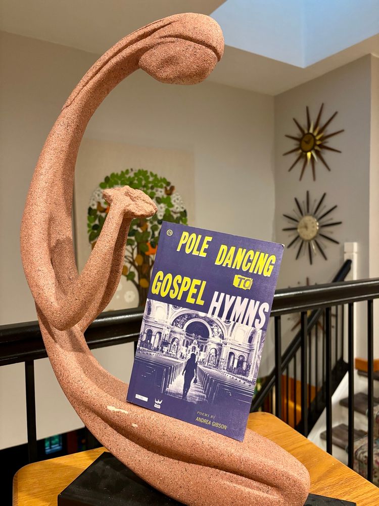 A copy of Andrea Gibson’s Pole Dancing to Gospel Hymns, sitting in a statue that is looking down at the flower of poesy it holds, foregrounded while the clocks and the tree of life in the background are blurred.