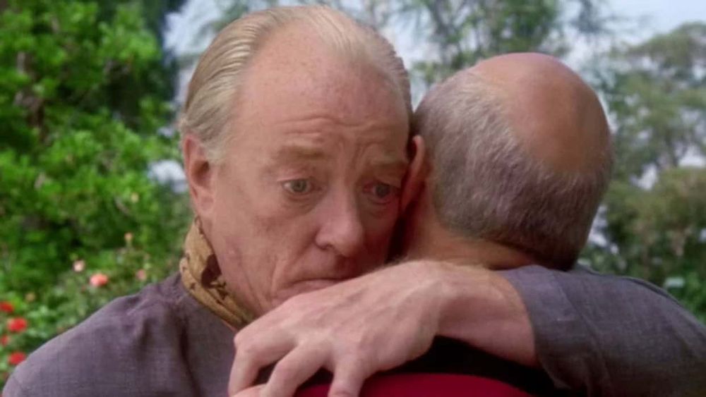 An older man balding hugs another older balding man.