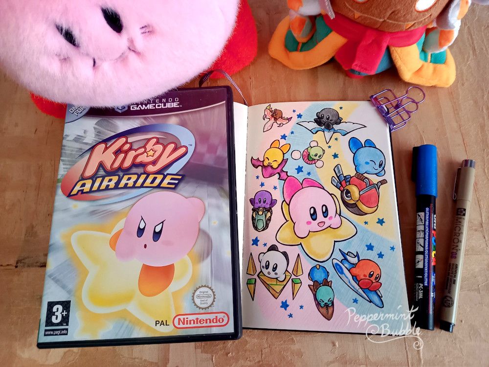 A photo of my sketchbook laid open, with a colorful sketchy colored pencil drawing with black ink lineart of 10 differently colored Kirbys each riding a different air machine from Kirby Air Ride, flying down along a pastel-colored rainbow. My copy of Kirby Air Ride for the Gamecube is placed next to the sketchbook, and my memetic fluffy classic Kirby plushie and Taranza plushie can be seen at the top of the photo, which has been staged on my work desk on top of my wooden painting surface.