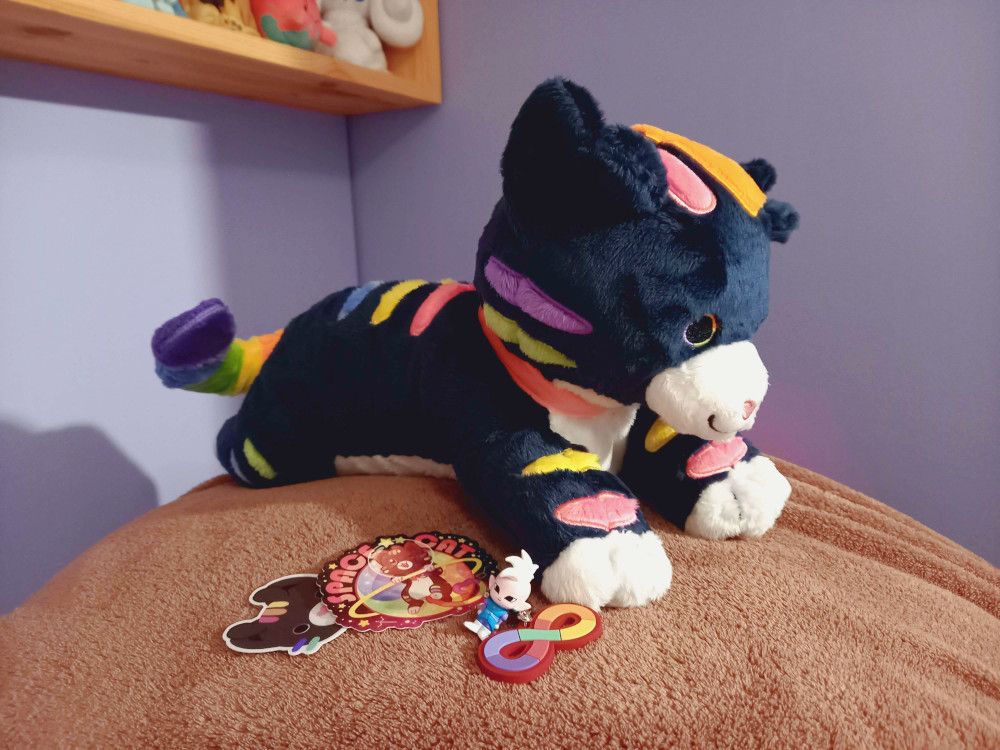 A photo of my Critter the space cat plushie, designed by Hannimations. The plush is on top of a brown blanket, and I've put Critter stickers, the autism pride symbol charm that belongs on Critter's neck, and my bath bomb Shin minifigure next to its paw.