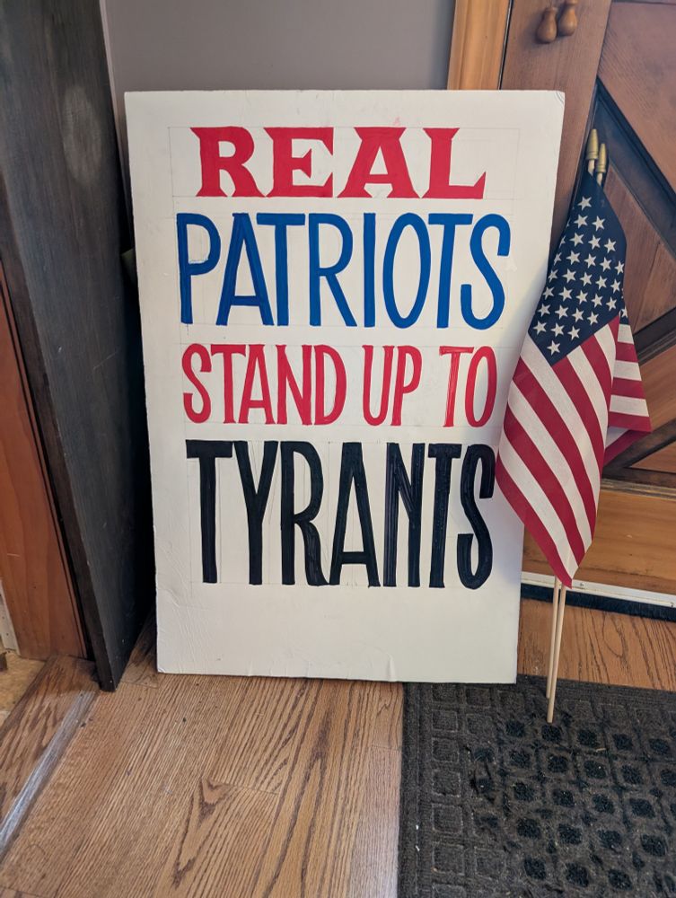 A poster with the words "Real Patriots Stand Up To Tyrants" with a small American flag leaning next to it