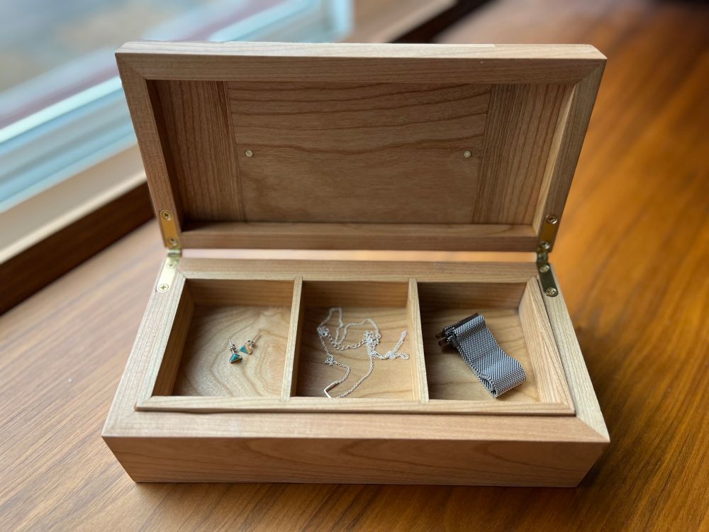 A wooden box with the lid open. There are 3 small sections inside; each holding a piece of jewelry. 