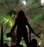 The silhouette of a person stood with their arms out and down to the sides. The person has long hair and is holding a top hat in their left hand and a stick in their right. Behind the person is a large machine resembling a wheel with spokes attached to a stand. Light from fireworks make the background green and smoky.