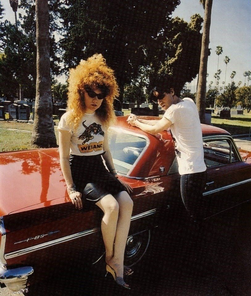 Poison ivy from the cramps sitting on a red oldsmobile looking cool as fuck with big hair and lix in the background