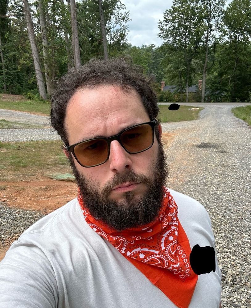 Selfie of a white guy with dark brown hair and beard, wearing brown tint sunglasses, a light grey shirt, and an orange bandana tied around his neck like a cowboy. He’s making a serious face. 