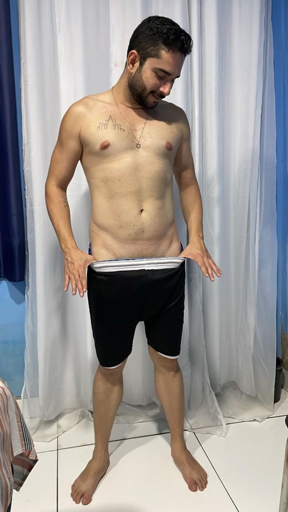 Imagem 2: Mesmo homem da imagem anterior, agora de frente e sorrindo levemente enquanto olha para dentro do short preto com elástico branco, que ele puxa com ambas as mãos. Ele continua sem camisa e descalço, em um ambiente com piso branco e cortinas claras ao fundo.