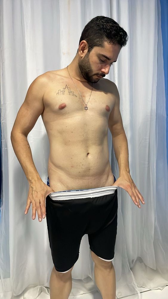 Imagem 1: Homem em pé, de perfil levemente inclinado para frente, olhando para dentro de seu short preto com elástico branco que está sendo puxado com as duas mãos. Ele está sem camisa, com colar de estrela de Davi no pescoço e uma tatuagem minimalista de prédios no lado esquerdo do peito. O fundo é composto por cortinas brancas e azuis.