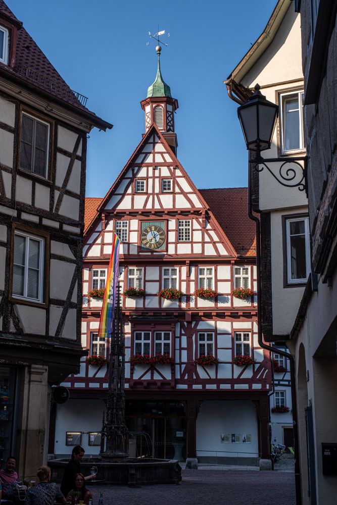 In einer Altstadt mit Fachwerkhäusern blickt durch eine Gasse auf ein Fachwerkhaus mit großer Uhr, spitz zulaufenden Giebeln und einem Turm. Vor den Fenstern sind Blumenkästen, am Haus hängt eine Regenbogenfahne.