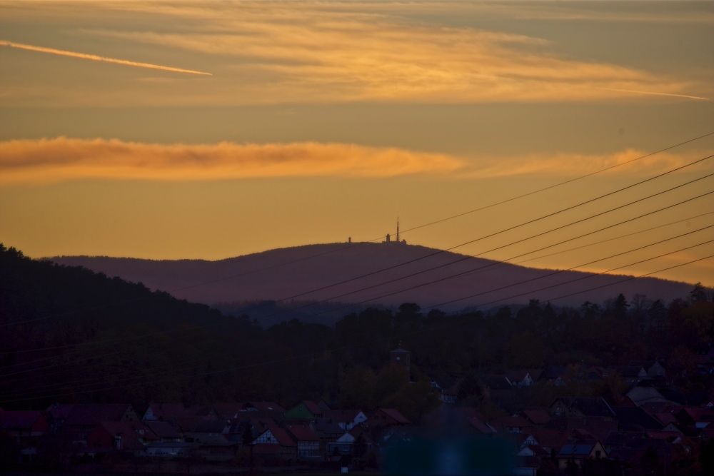 Silhouette des Brockens im Harz mit Sendemasten auf dem Gipfel bei Sonnenuntergang, darunter ein Dorf mit Häusern und Stromleitungen.