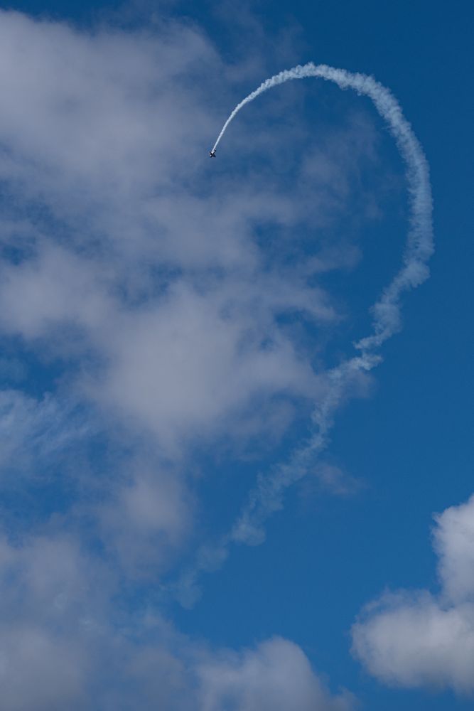 Ein kleines Flugzeug, ein Doppeldecker hoch oben am Himmel, ein Kunstflieger ist dabei, eine Figur mit Überschall zu fliegen. Eine Rauchschwade markiert die Flugbahn, die hoch und in einem Bogen nach unten führt.