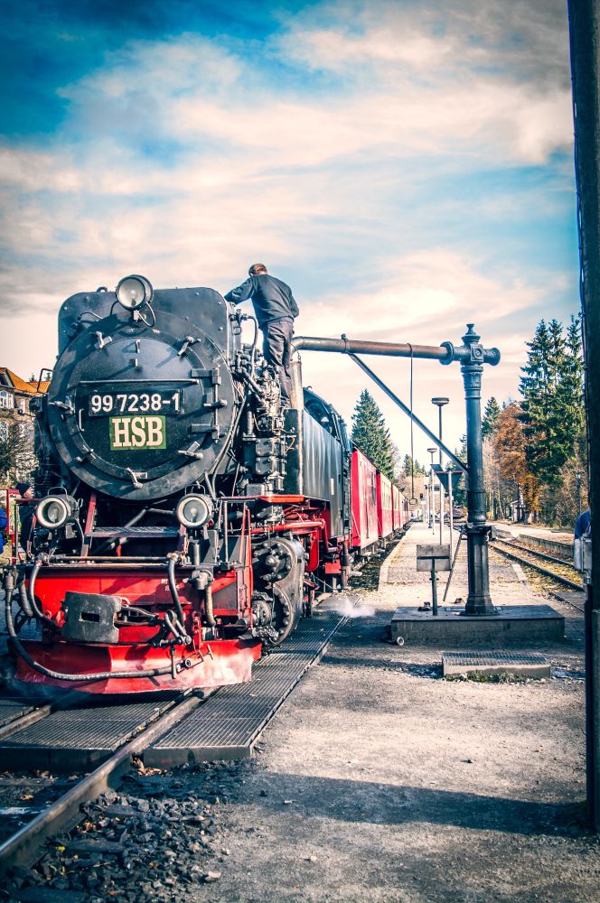 Ein Dampfzug mit roten Waggons steht in einem Bahnhof, er wird dir ein schwenkbares Wasserrohr mit Wasser gefüllt, der Heizer steht oben auf der Lock. An der Dampflock steht vorne eine Zahlenreihe und drei Buchstaben, HSB.