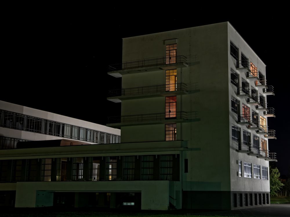 Bei Nacht. Ein modernes Gebäude, was über Zwischenbauten mit anderen Gebäuden verbunden ist. Es ist ein schlichter kantiger Bau mit einfachen Balkonen. Die Fenster sind teilweise erleuchtet, man schaut schräg auf das Gebäude.