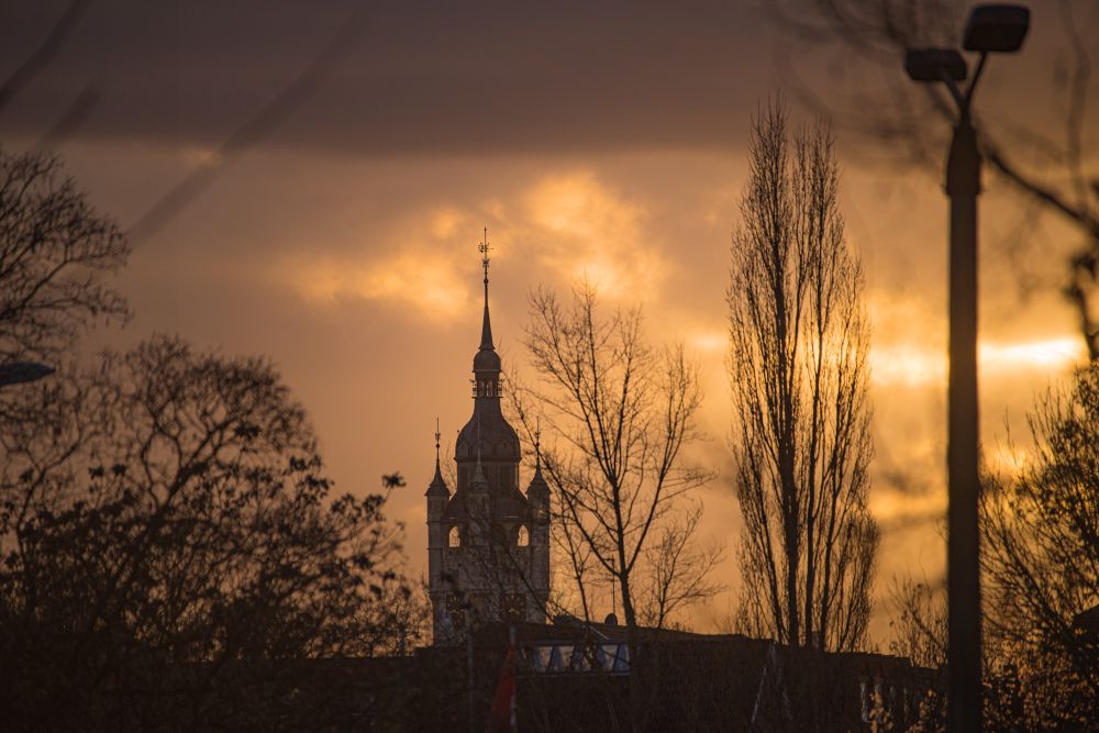 Im Winter die Bäume sind laubfrei, der Blick geht an den Bäumen vorbei auf einen großen Turm, an dem kleinere Türme sind. Die Turmspitze ragt sehr hoch. Der Himmel ist bewölkt und vom Sonnenuntergang golden eingefärbt.