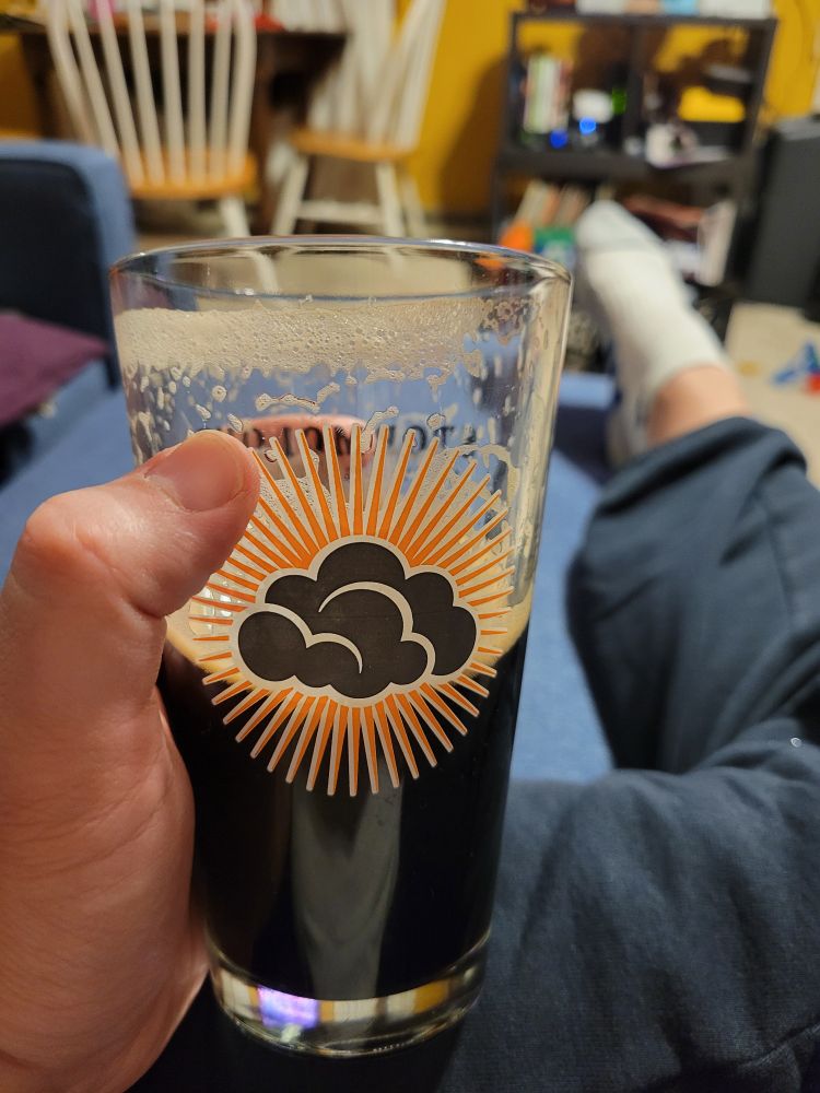 Photo of my left hand holding a Stormcloud Brewing Co. pint glass halfway full of Bell's Kalamazoo Stout; the photo also shows my right leg extended on a blue ottoman