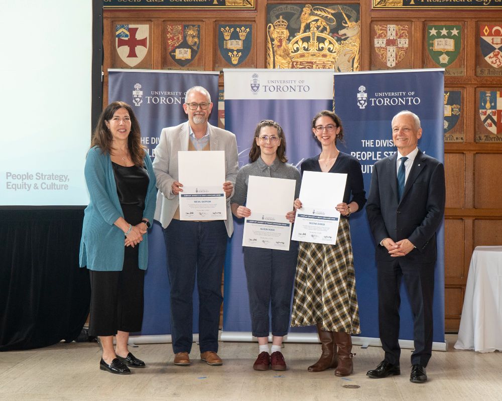 TATP staff hold their awards, standing in Hart House Great Hall with President Meric Gertler