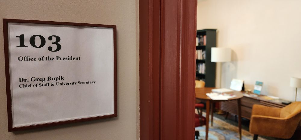 A photo of a door sign stating Greg's title chief of staff and university Secretary with open door to his office to the right 