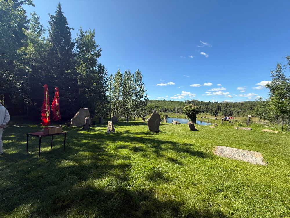 Photo of a standing stone circle in a grassy meadow with aspen trees around. There are two banners with ogham writing on them visible at the entrance to the circle. 