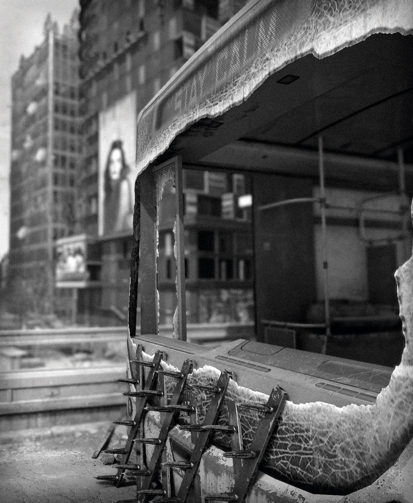 Dying Light 2: scenery shot in b&w; close up of a bus with shattered windows and the text "Stay Calm" at the top and bloody spikes attached to its front. The city can be seen behind it; two tall buildings with billboards (blurred) of which one appears to show a woman.