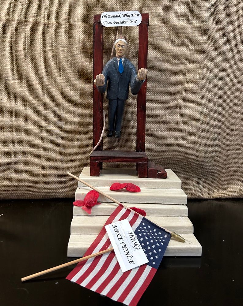 A sculpture of Mike Pence hangs from a gallows at the top of a set of marble stairs. Red hats litter the stairs. Also on the stairs are a fallen American flag and a sign reading “Hang Mike Pence.” Pence has his hands out as if pleading or praying. At the top of the gallows is a sign reading “Oh Donald, why has thou forsaken me?”