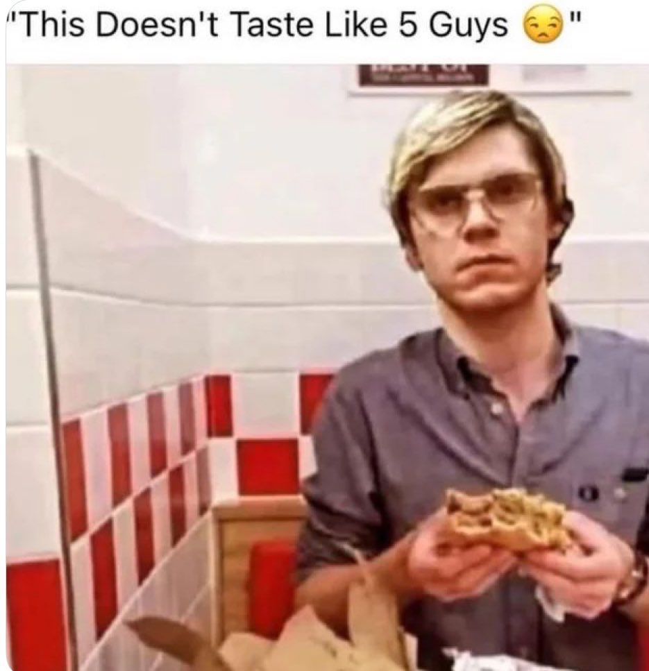 A young man with blond hair and glasses sits in a fast food booth with red and white checker tiles. He wears a gray button up shirt and holds a messy burger with both hands, staring blankly ahead. On the table are a brown paper bag and crumpled foil. Meme style top text reads This Doesnt Taste Like 5 Guys with a disappointed face emoji.