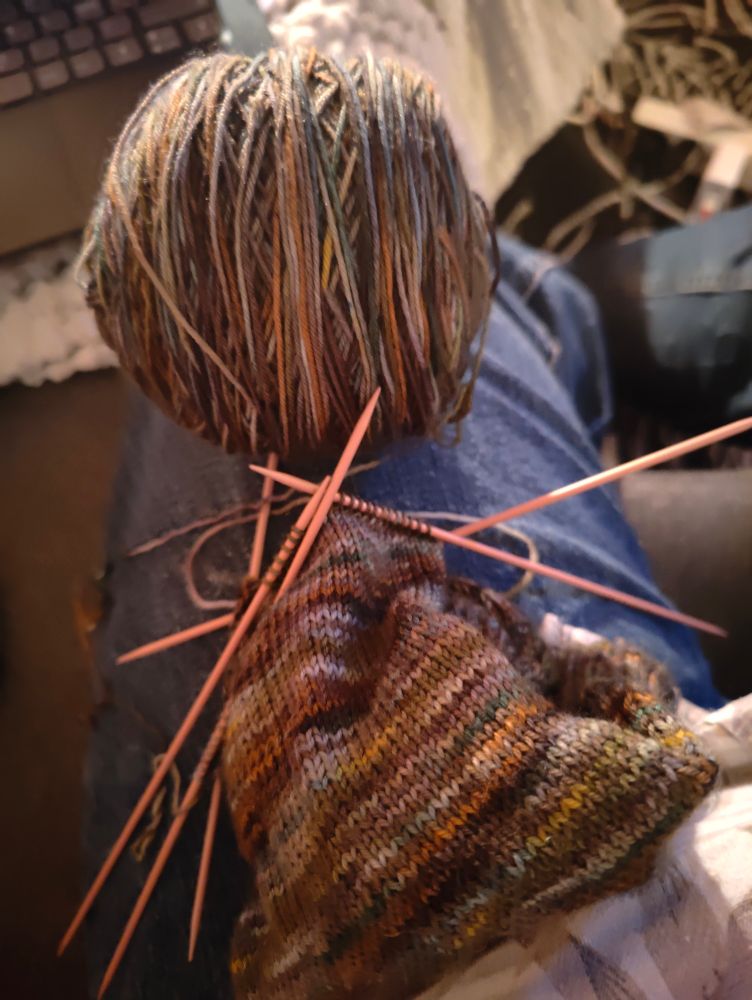 Earthy toned sock project on knitting needles, yarn ball next to the project. Dim lighting giving a warm feel to the photo.