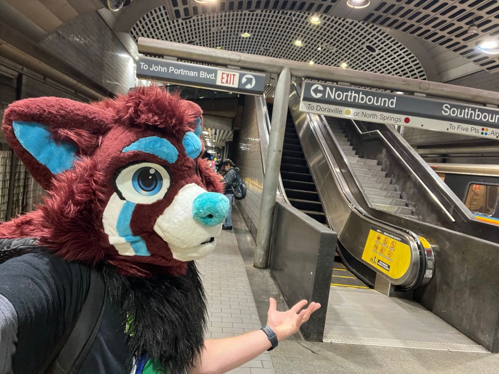 A red pana fursuiter standing in a subway station.