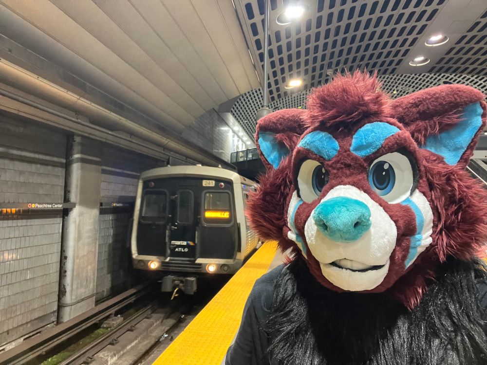 A red pana fursuiter standing next to a subway train. 