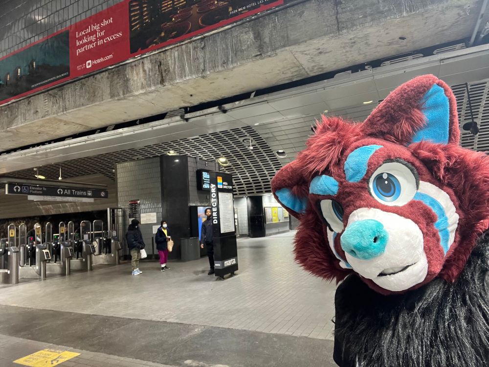 A red pana fursuiter standing in a subway station.