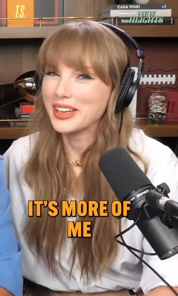 Taylor Swift with her hair down in a white shirt with headphones on talking into a podcast mic with “it’s more of me” written on the screen as captions to what she was saying