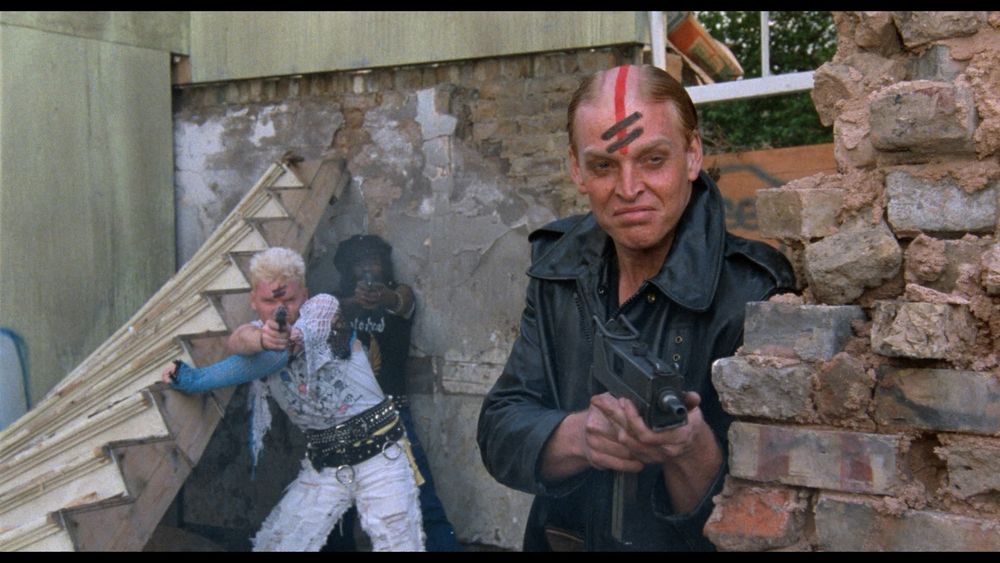 A skinhead with a red line vertically down his forehead with 2 diagonal black slashes above the eyebrow, wearing a black leather jacket and holding an Uzi, stands next to a crumbling brick wall in front of a man in a ripped white pantsuit and blue cast, leaning against a fallen staircase