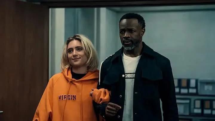 A short blonde girl in an orange hoodie walks arm in arm with a Black man with a salt & pepper goatee, wearing a black jacket and white t-shirt