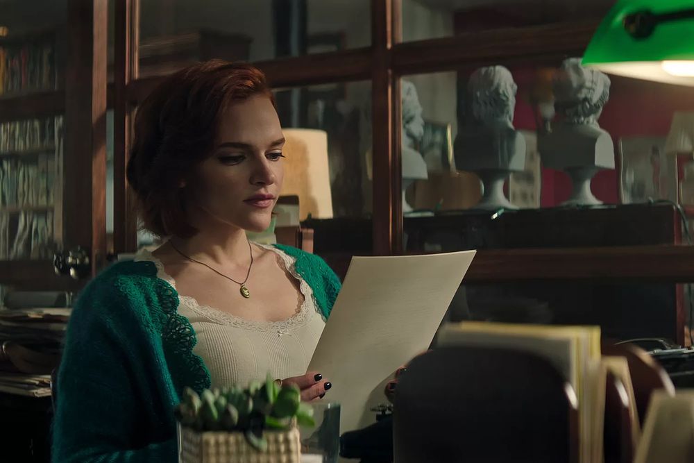 A red head wearing a white camisole and green sweater reads a piece of paper in an office