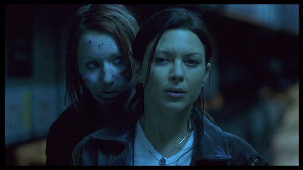 A pale faced ghost peers over the left shoulder of a dark haired woman in a black leather jacket standing on a subway platform