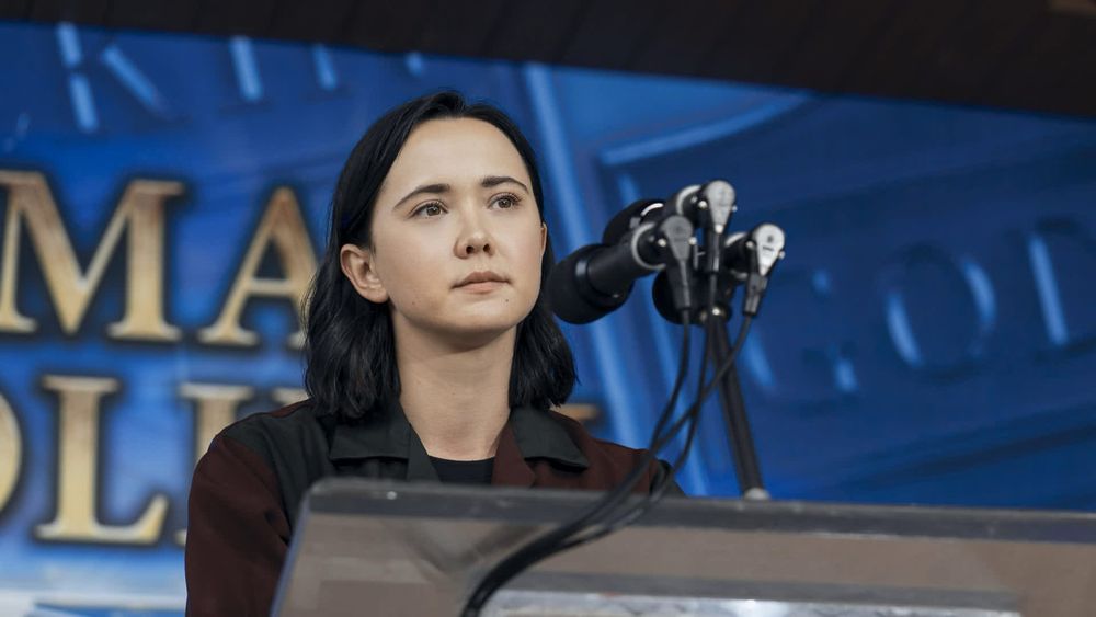 A dark haired young woman stands pensively in front of a microphone