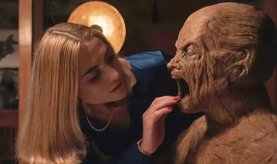 A blonde woman in a blue dress leans down to stick her fingers in the mouth of a wooden man