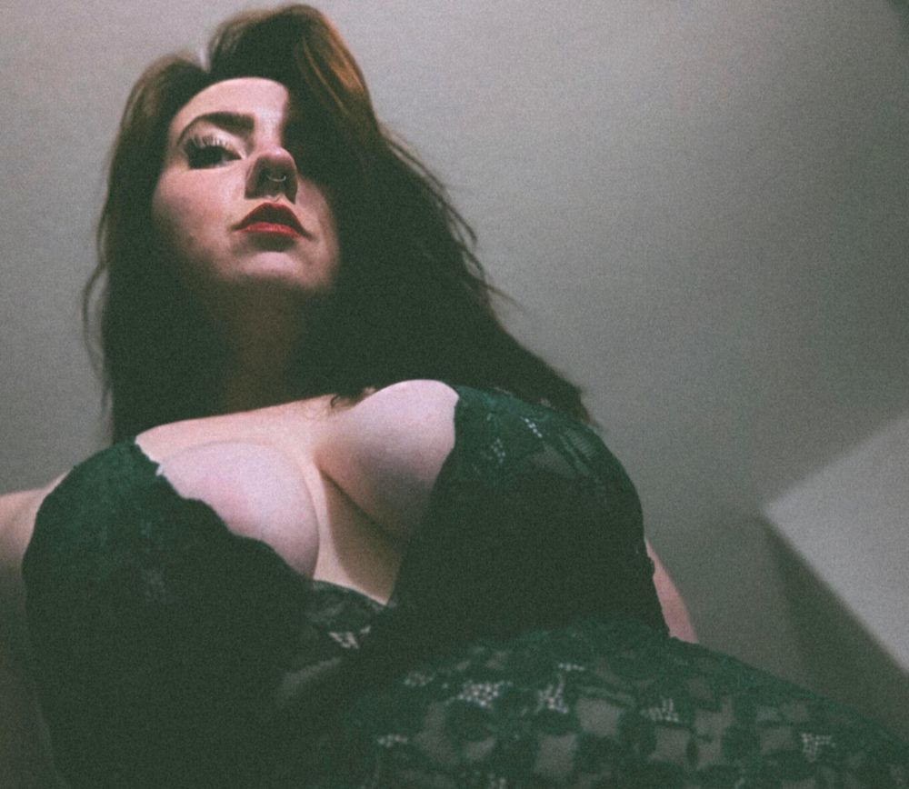 Grainy image of Lenore sins leaning over a camera in a lace emerald bodysuit 