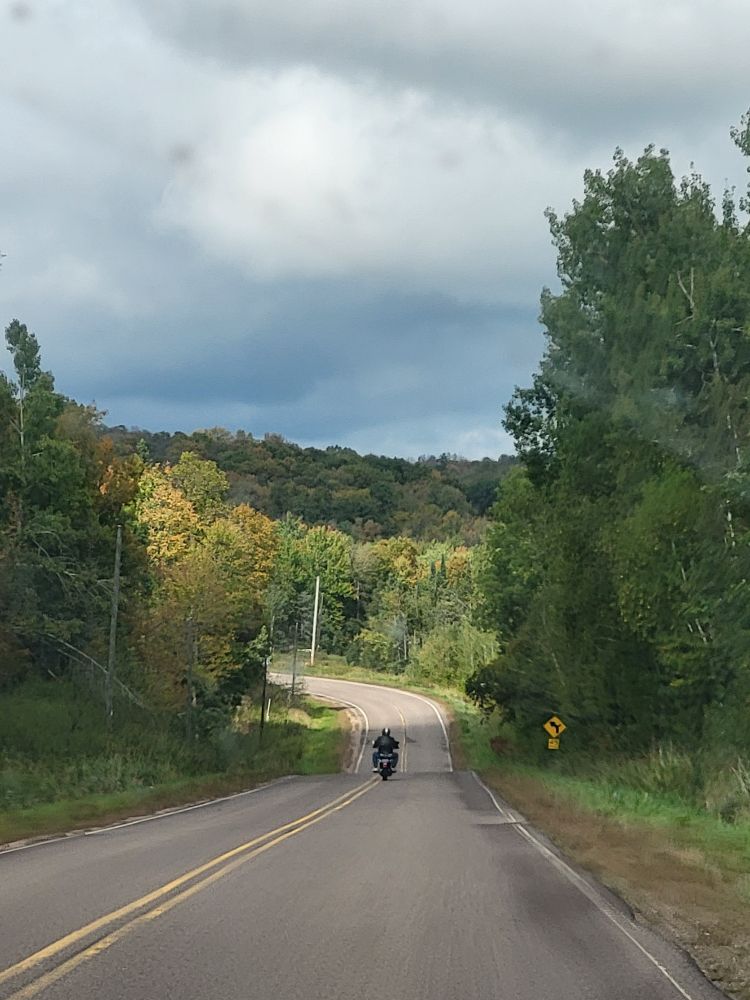 View of the road ahead, bending to the left.  A view of the tree covered hills is straight ahead, not too far back from the bend in the road.  Trees line the road on either side.  Ahead on the road is a (stranger) guy on a motorcycle.  He really adds to the composition, and his license plate is far blurry enough i don't have to worry about him being doxxed.