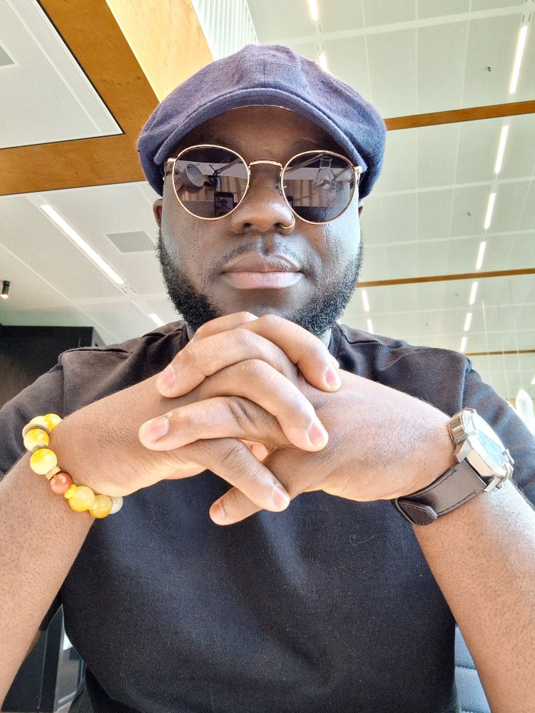 A Black man sitting indoors with fingers interlaced in front of him, wearing a navy flat cap, round sunglasses, a nose ring, a black shirt, a yellow bead bracelet, and a dark brown leather wrist watch. He has a short beard and a calm but confident expression, as he looks into the camera.