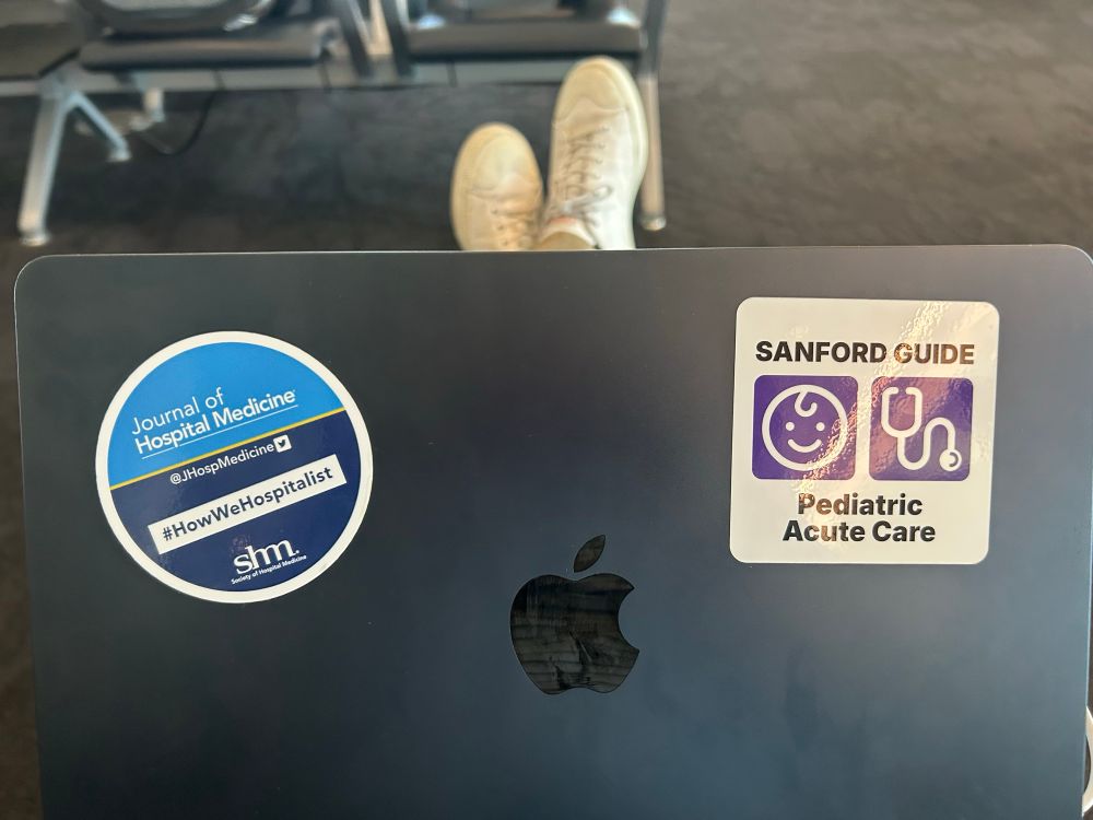 Photo of a navy blue Mac laptop with 2 stickers slightly above and lateral to the Apple logo. On the left is a circular Journal of Hospital Medicine-branded sticker. The top half of the circle is a light blue, the bottom half is dark blue and a thin yellow line bisects the two halves. The top half has the Journal of Hospital Medicine logo in white and the bottom half has, also in white, #HowWeHospitalist and the society of hospital medicine logo. The image on the right is a white square with rounded edges. It resembles how app me appear on phones and tablets. In black lettering are the words “Sanford Guide” at the top and “Pediatric Acute Care” at the bottom. Centered in between are two smaller squares in purple with images in white. The left has a stick figure baby head and the right is a stethoscope; these represent the Sanford Guide to Pediatric Acute Care logo. 