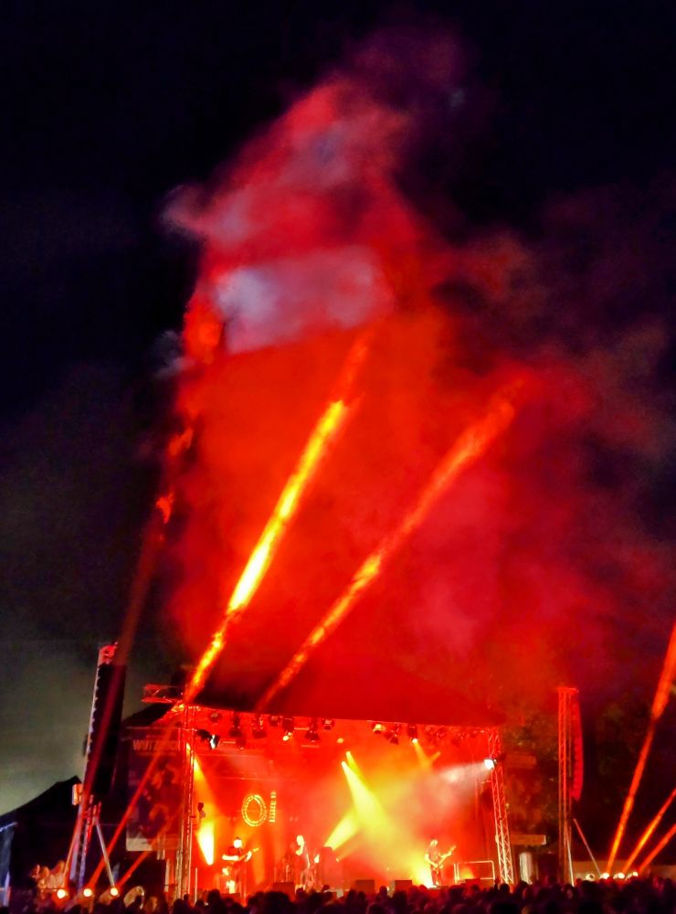 Eine mittelgroße Festivalbühne auf einem Festival, es ist dunkel, Strahler, rotes Licht und Rauch. Im Hintergrund ein rot leuchtends "Oi!" An der Bühnenrückwand. Auf der Bühne Oidorno, vor der Bühne schemenhaft Menschen. Es sieht sehr voll aus. 