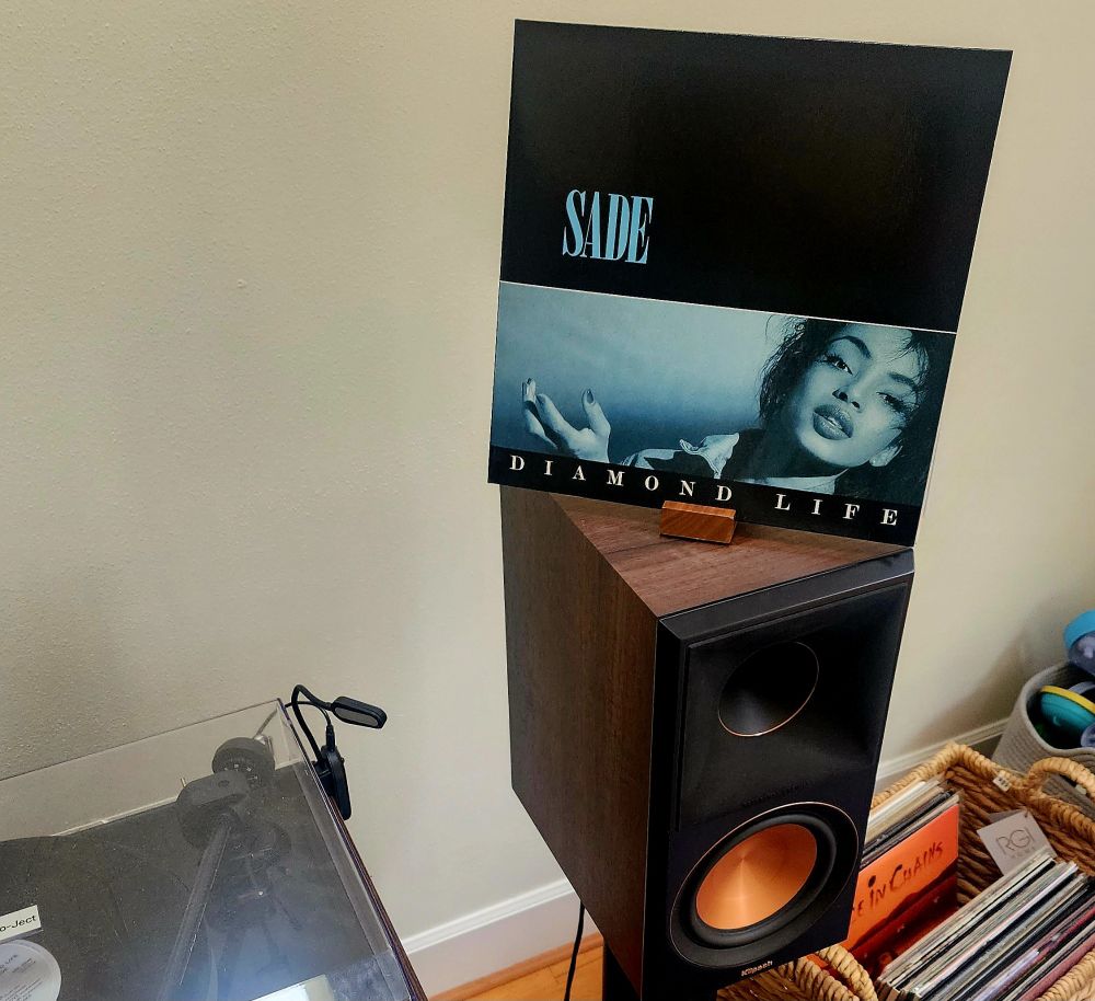A copy of the album cover atop a speaker. A basket of records sits to the right and a turntable stands to the left. 
