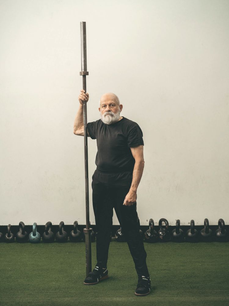 Older weightlifter holding bar vertical in front of kettle bells in gym