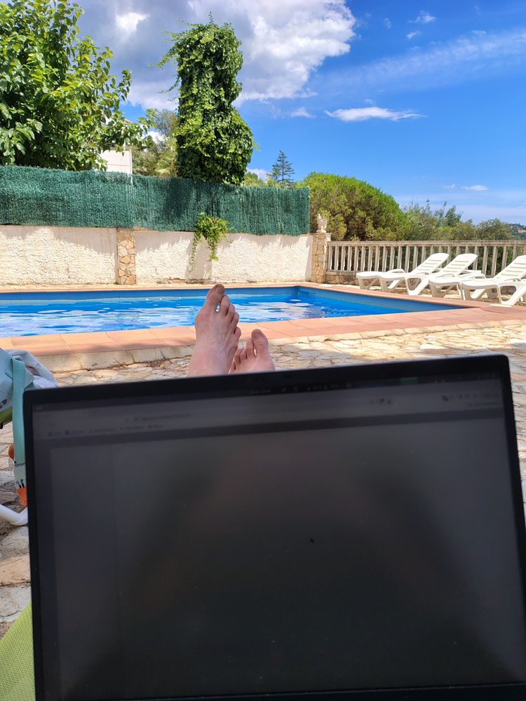 My laptop and feet as I lie on a sunbed beside a swimming pool.