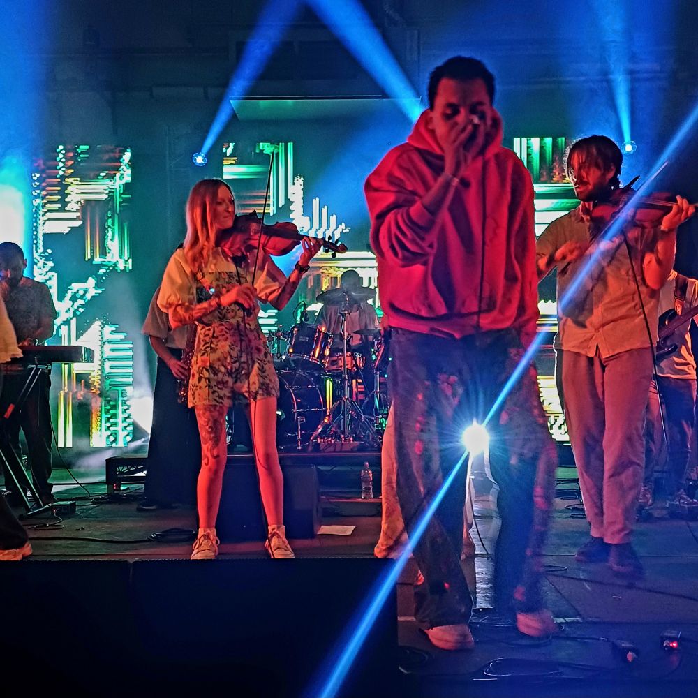 Photo of the Tinderbox Orchestra on stage with a young man in a red hoodie and embroidered jeans rapping into a microphone while two young violinists in casual street clothes play, with drums and colourful digital screens in the background and a blue laser light catching the camera.