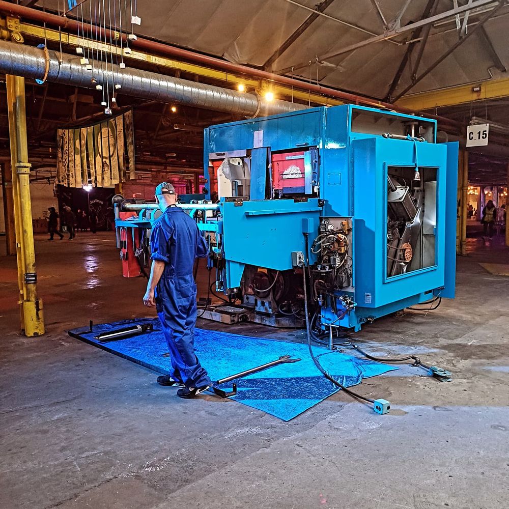 A photo of the Paper Factory floor, with grey concrete and industrial yellow metal beams in the background, a blue industrial tarpaulin on the floor, and a man who is a percussionist in a blue boilersuit with rolled up sleeves and a cap, drumming on a large square piece of blue machinery with wires coming out of it. This was part of a dance and percussion performance called 'Ghost in the machine' at Hidden Door Arts Festival.
